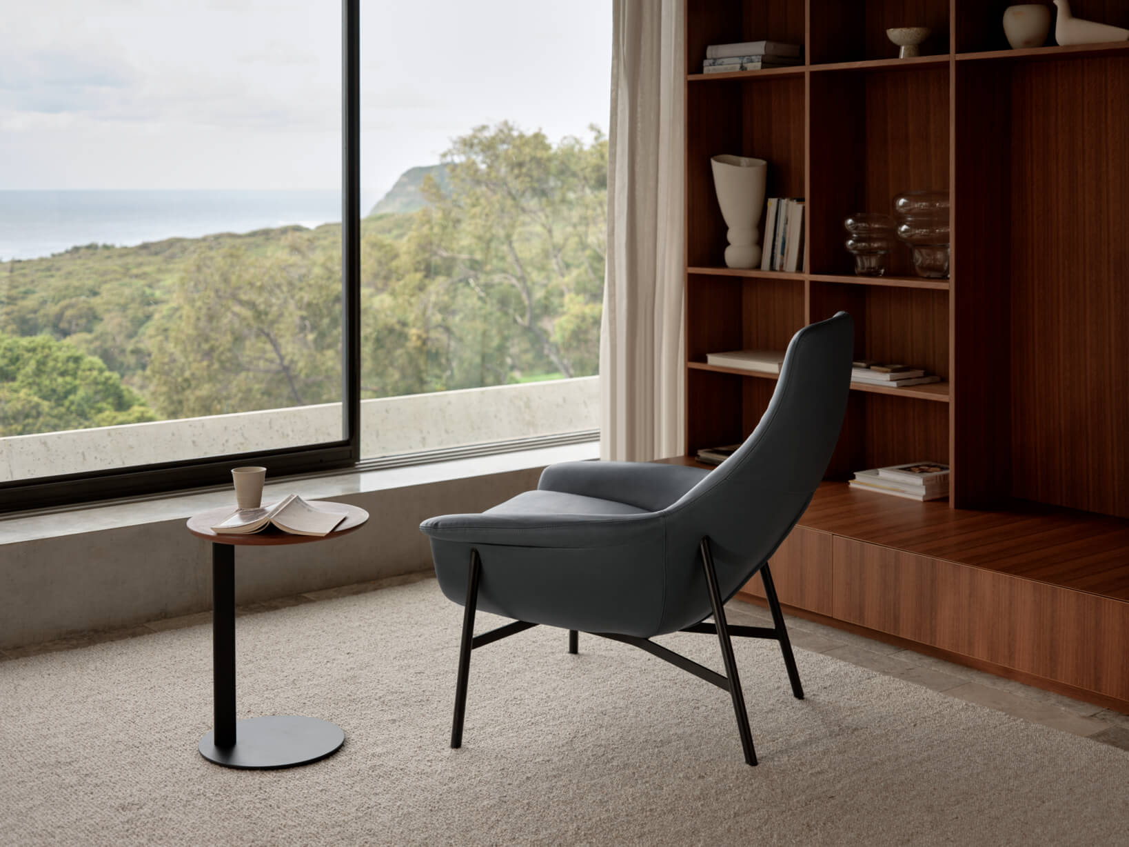 A refined leather armchair, paired with a timber side table, invites quiet moments of reading beside a panoramic coastal view.
