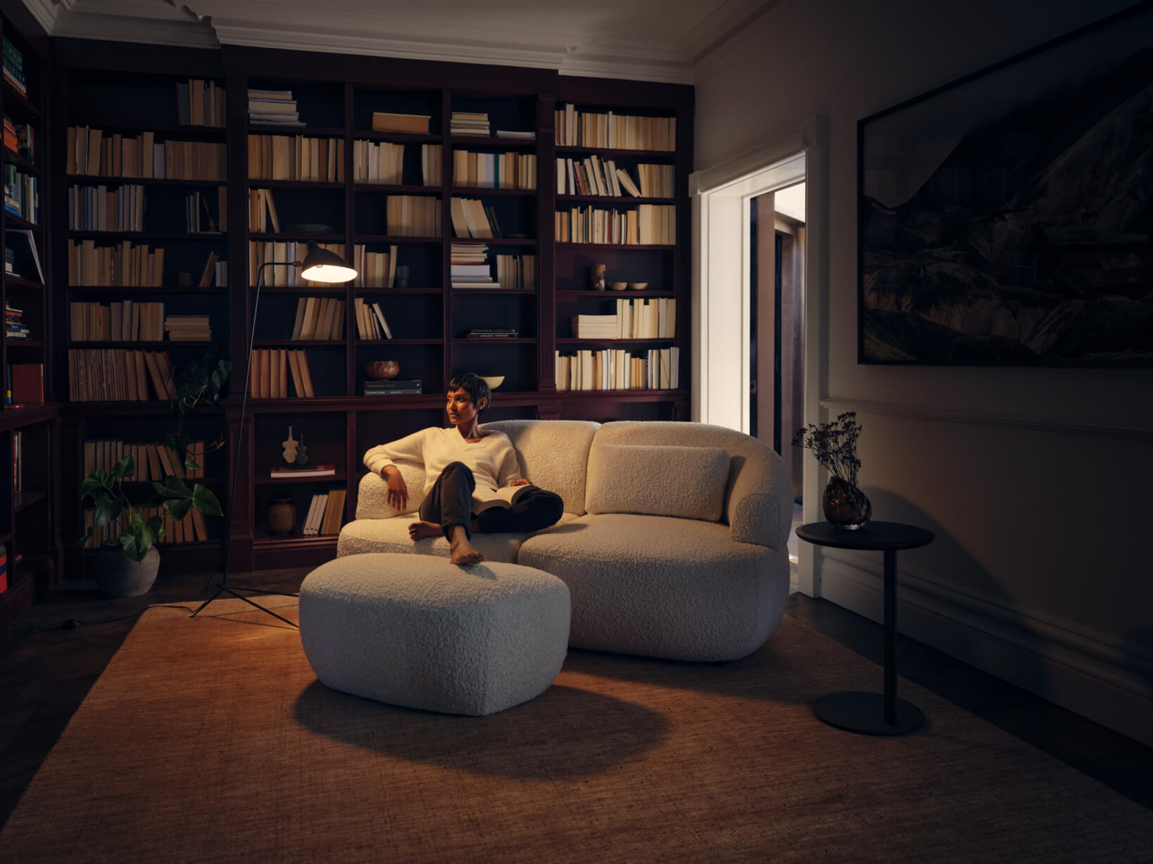 A woman lounges on a curved, beige, bouclé sofa paired with a matching ottoman in a warm-toned home library with bookshelves and ambient lighting.