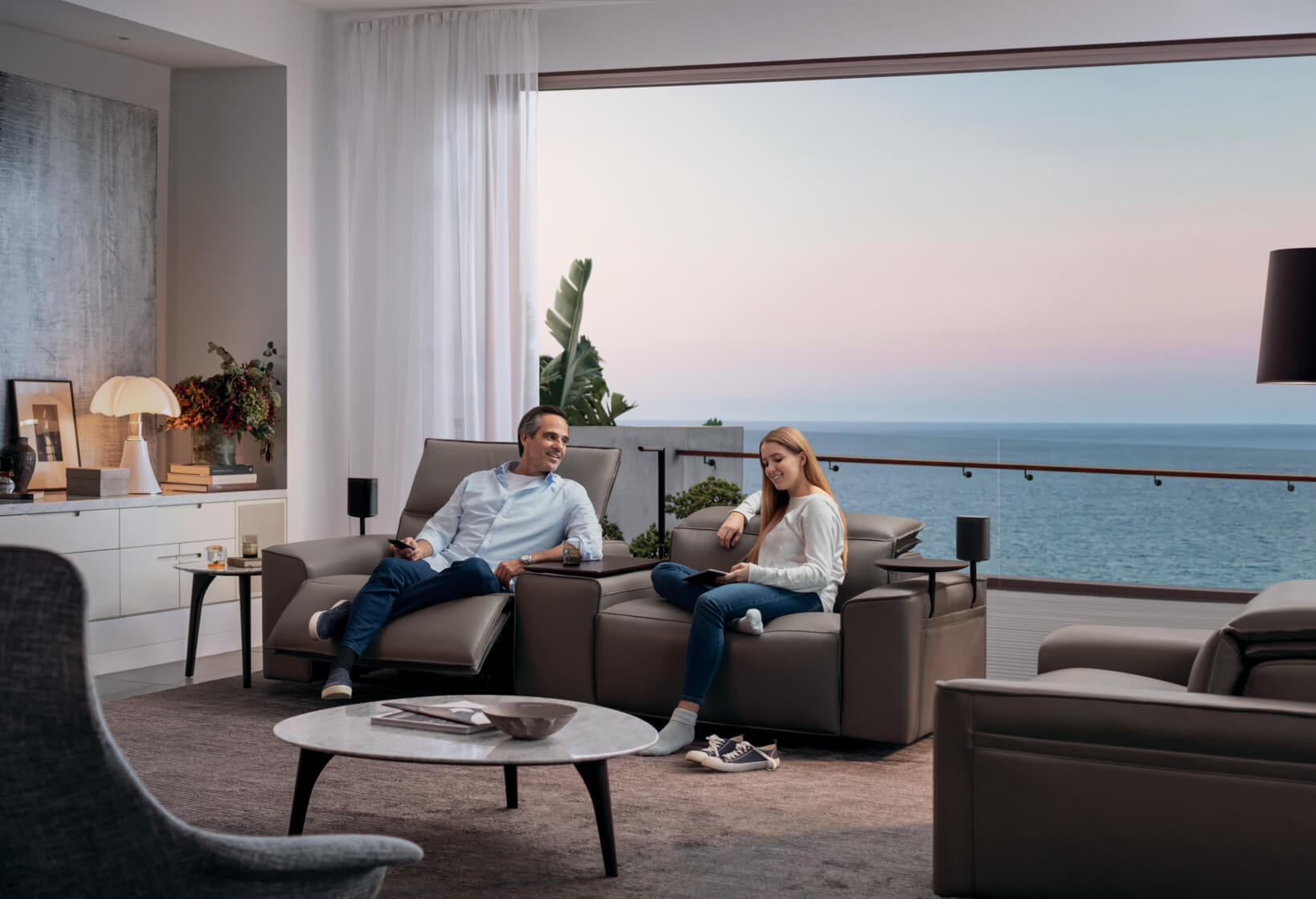 wo people lounging and reading on the King Cloud Recliner in a leather and timber finish, set against an expansive ocean-facing balcony.