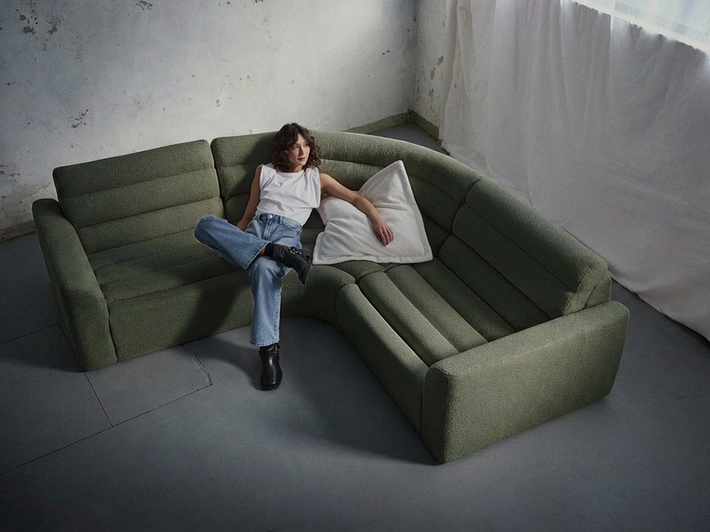 Woman reclining on a green curved modular sofa with high back cushions in a light concrete studio space.