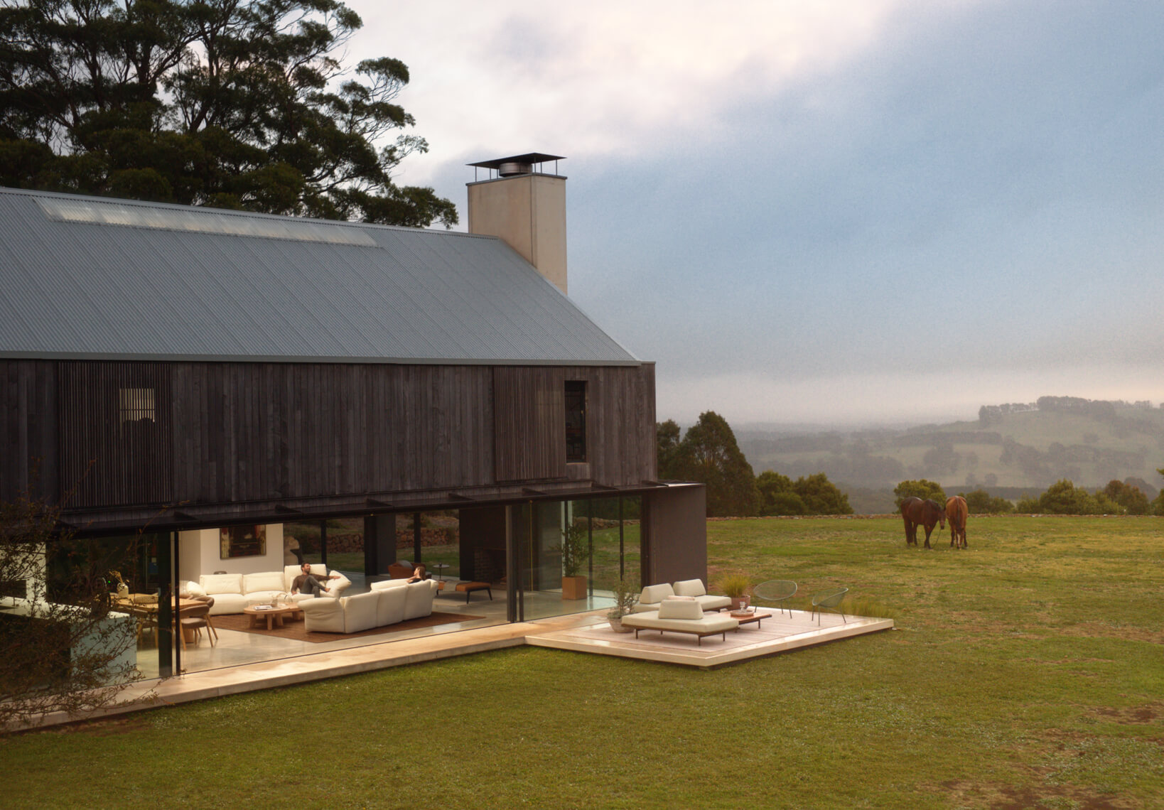 The exterior of rural home, modern farmhouse-style, with dark timber cladding opening to a terrace and outdoor lounge, overlooking fields.