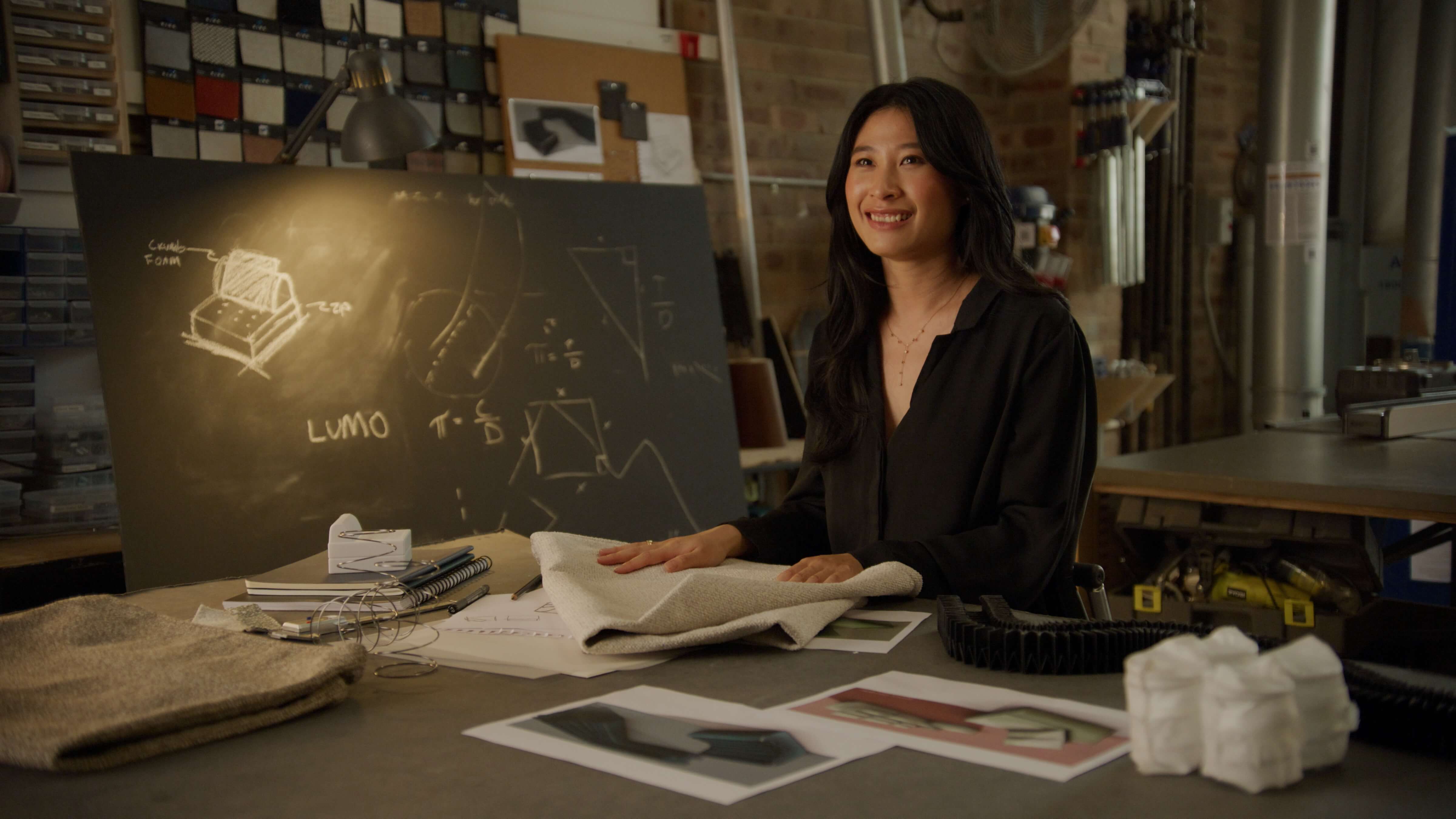 Designer sits in workshop beside blackboard sketches of sofas and sofas beds.