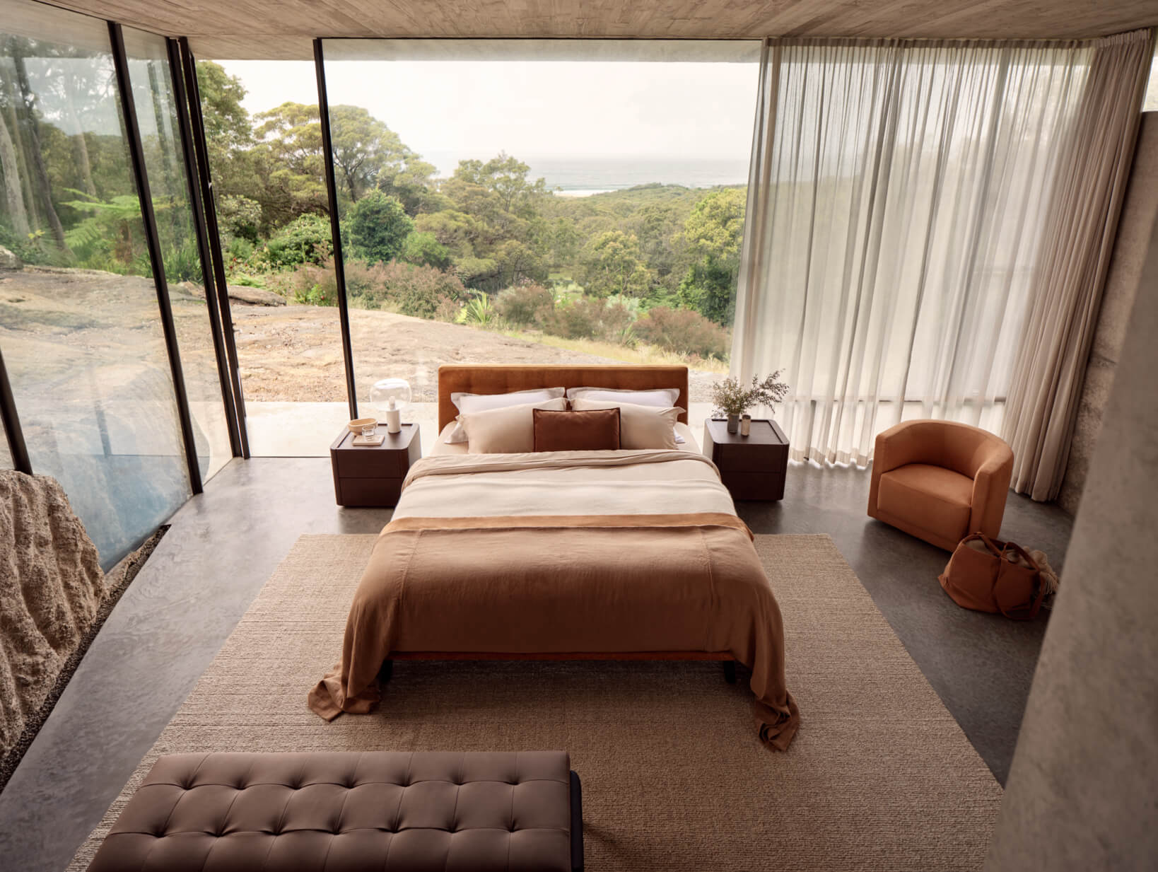 Master bedroom with bed, bedside tables, tufted bench at the foot, tub chair, ottoman, full-height windows with sheer drapery.
