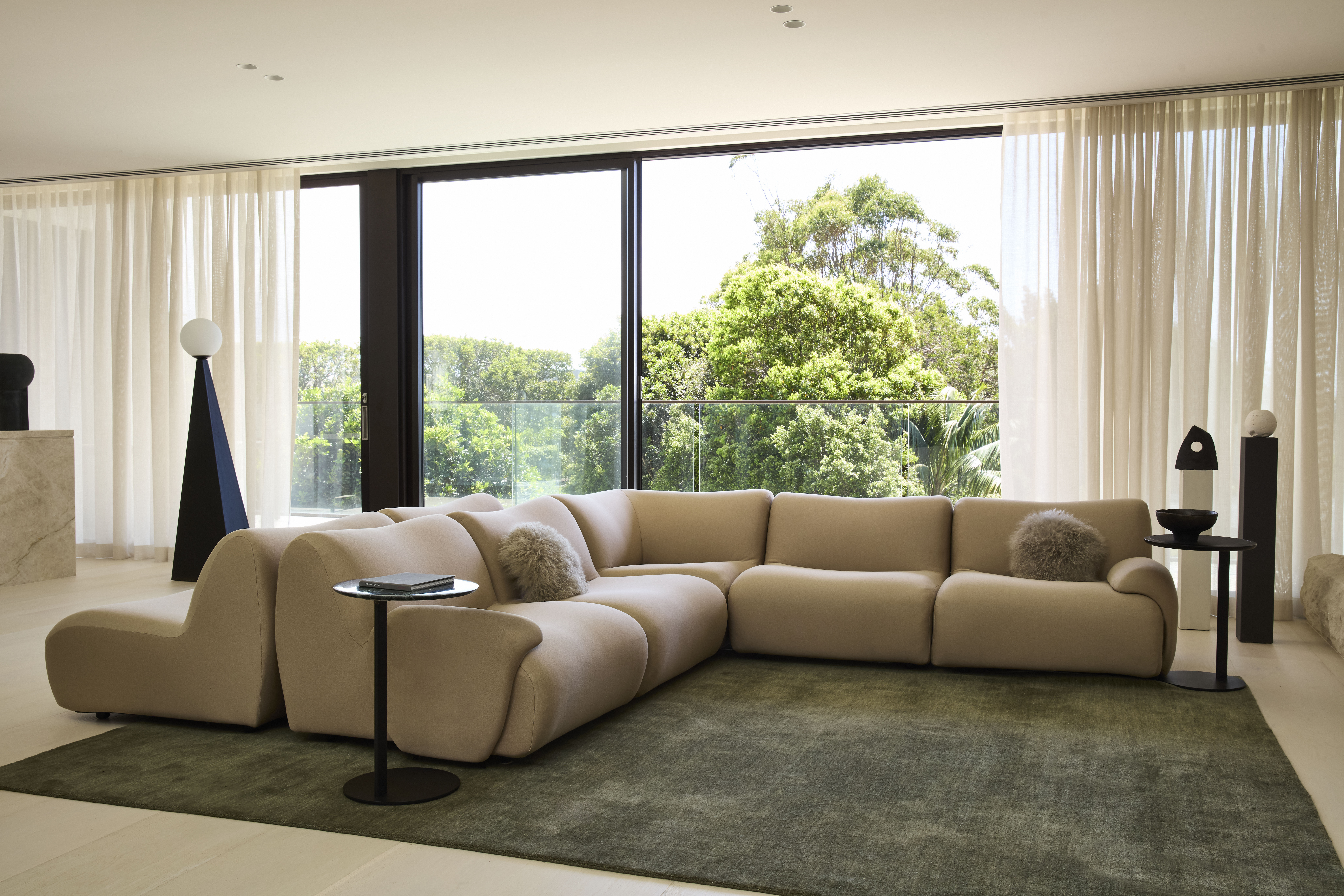 A wide shot of a light-filled living room bathed in earthy hues with a curved sofa in a muted sand colour, styled with a dusky green rug.