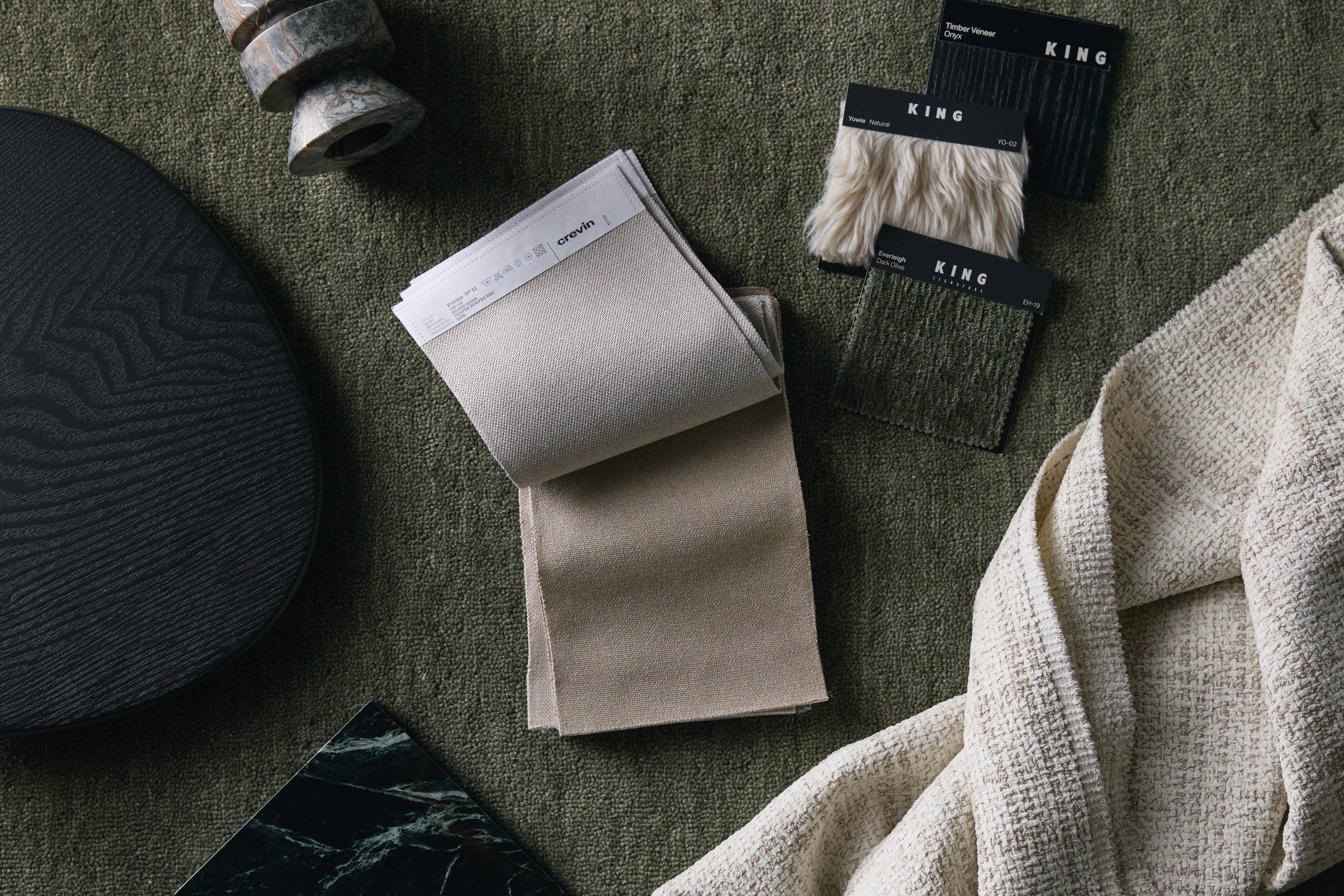 A flat lay of neutral and textured upholstery samples arranged on a deep green surface, with contrasting dark timber and marble accents.