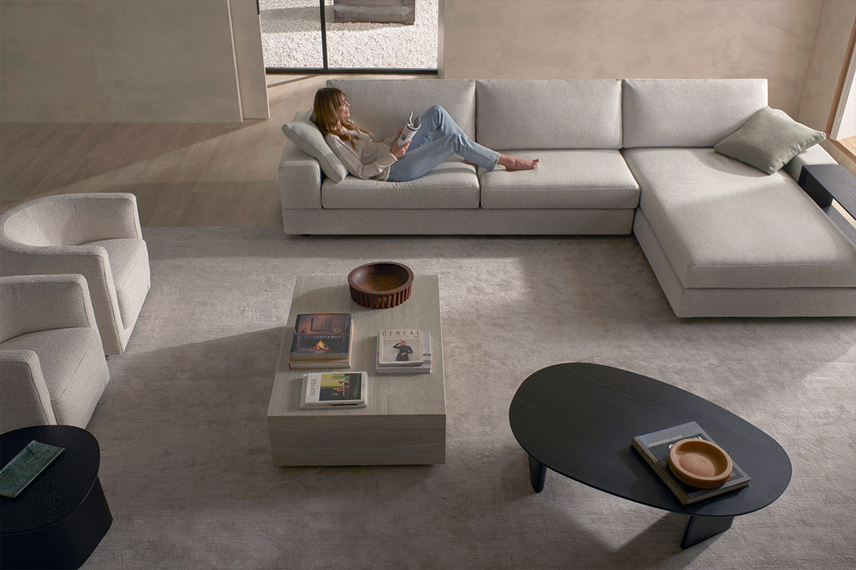 A minimalist living room with neutral furniture and a woman relaxing on a sofa.