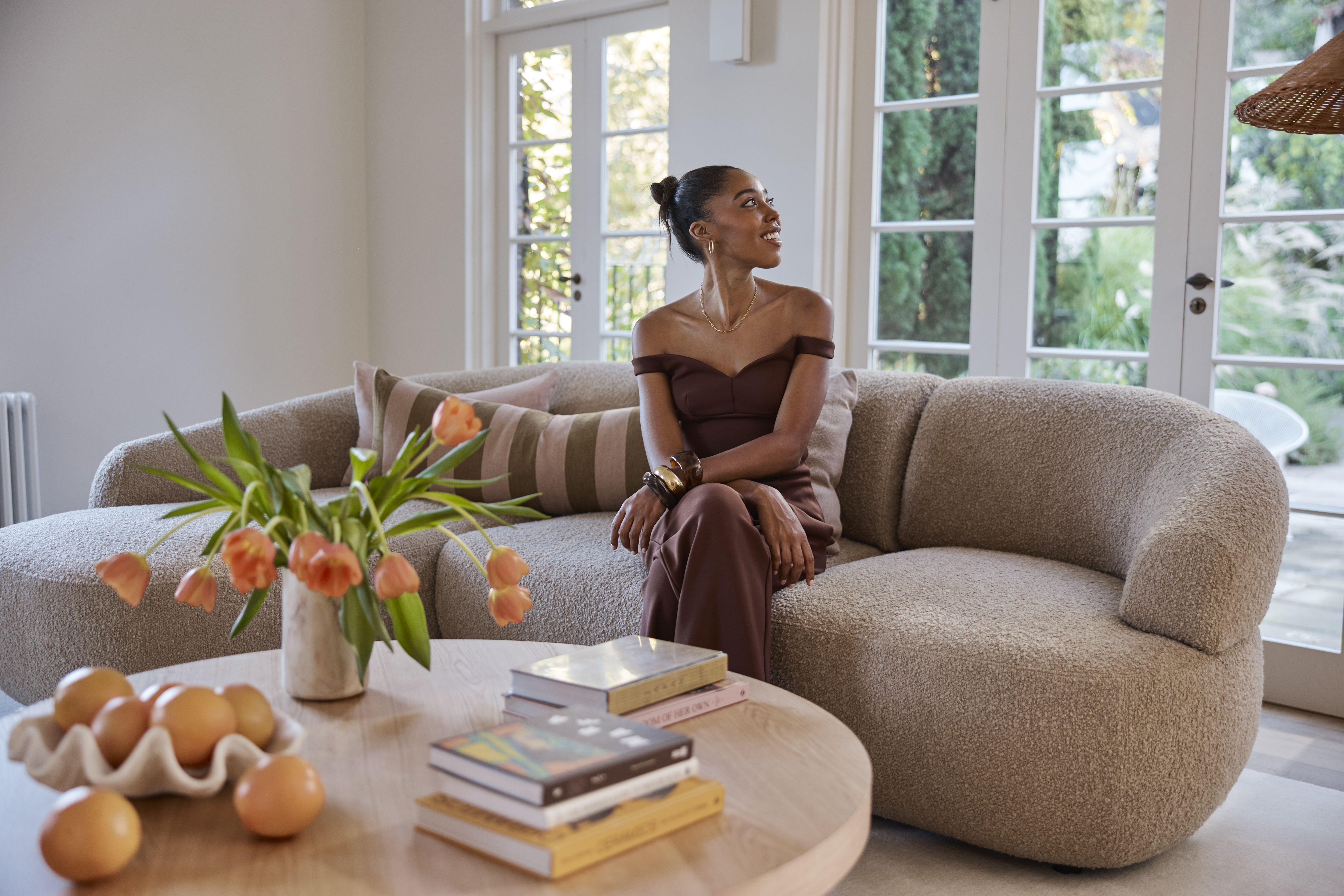 Woman in a brown dress sits on a curved beige sofa in a sunlit living room with soft, natural styling.