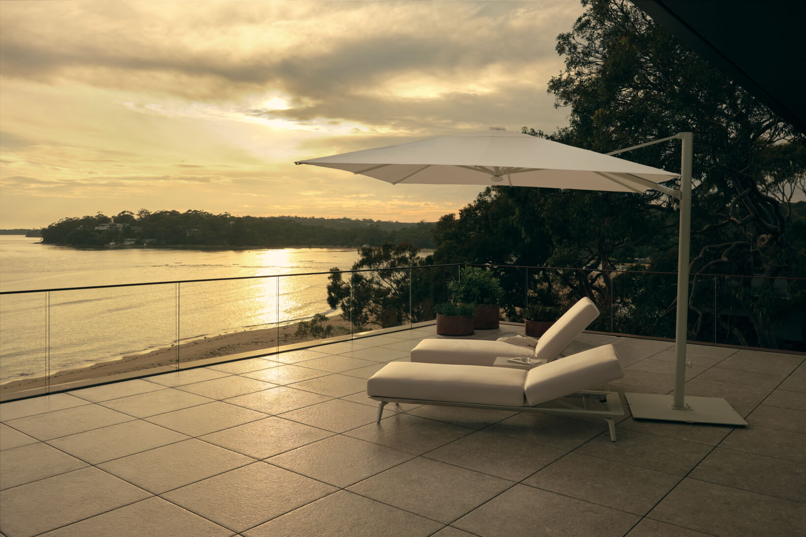 An outdoor tiled terrace with two white sun loungers and a large outdoor umbrella overlooking the water at sunset.