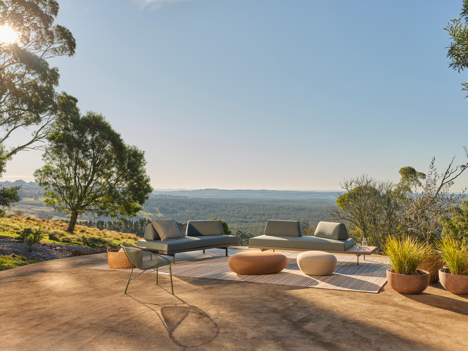 A patio with modular outdoor sofas, low organic ottomans, potted plants, and an outdoor rug, surrounded by a scenic valley.