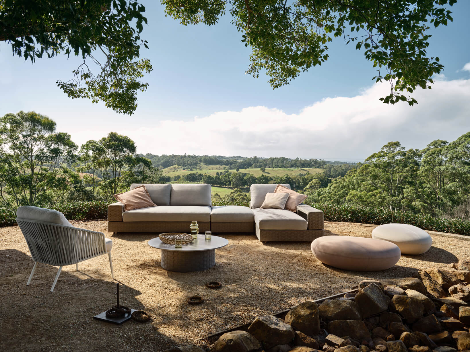 A lush green outdoor setting with a modular sofa, organic-shaped ottomans, a coffee table and occasional chair overlooking fields.