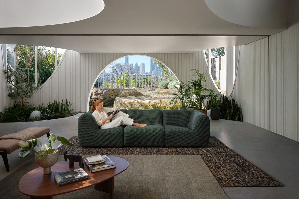 Modular sofa in a light-filled room with circular windows, layered rugs and timber coffee table overlooking a garden and city skyline.