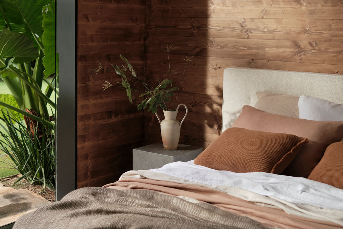 Earthy bedroom with timber walls, layered blush bedding and stone bedside table, opening to a lush garden view.