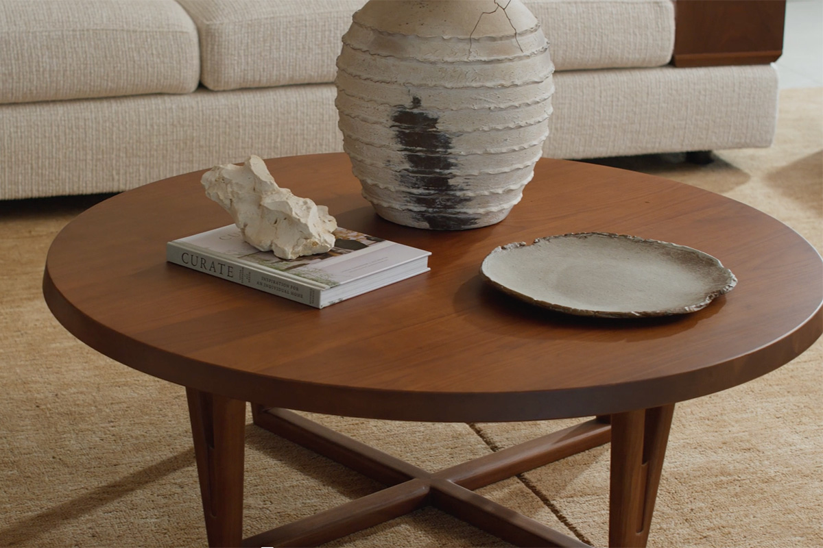 A round walnut coffee table styled with a tall sculptural vase and organic décor, with a neutral modular sofa in the background.