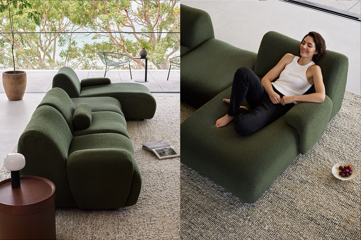 A woman lounges on a modular corner sofa with chaise finished in a deep green, boucle fabric, styled in a minimalist living room with lush views.