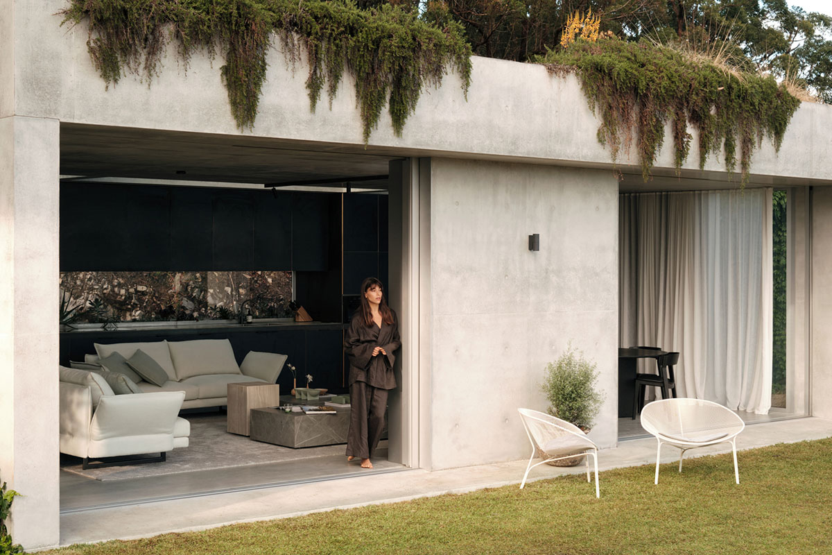 Concrete home with greenery above. Sliding doors open to reveal a neutral sofa in the living space, flowing seamlessly to the outdoors.