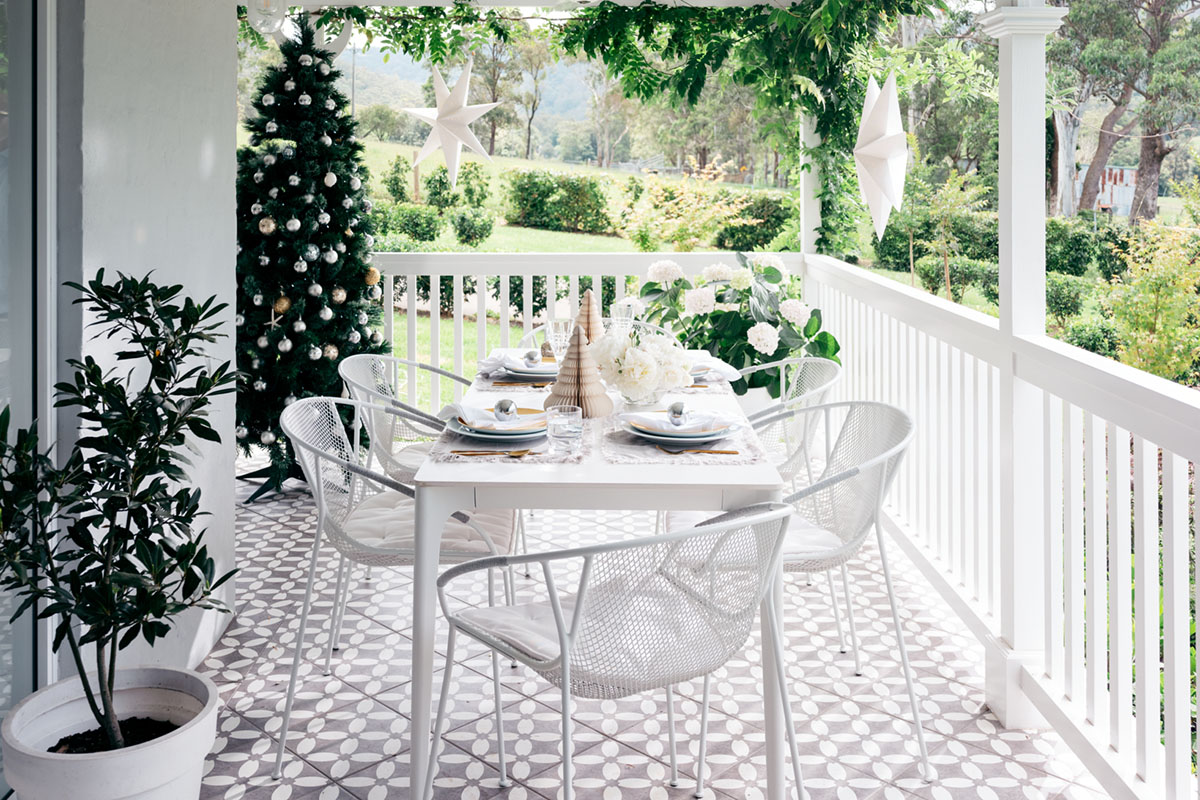 A veranda styled for celebration with white dining furniture, a Christmas tree, and silver festive decoration.