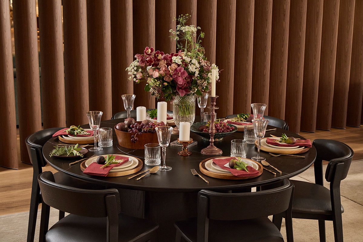 A round ceramic dining table with onyx timber dining chairs, styled abundantly with fruit, florals, and gold tableware for a festive celebration.