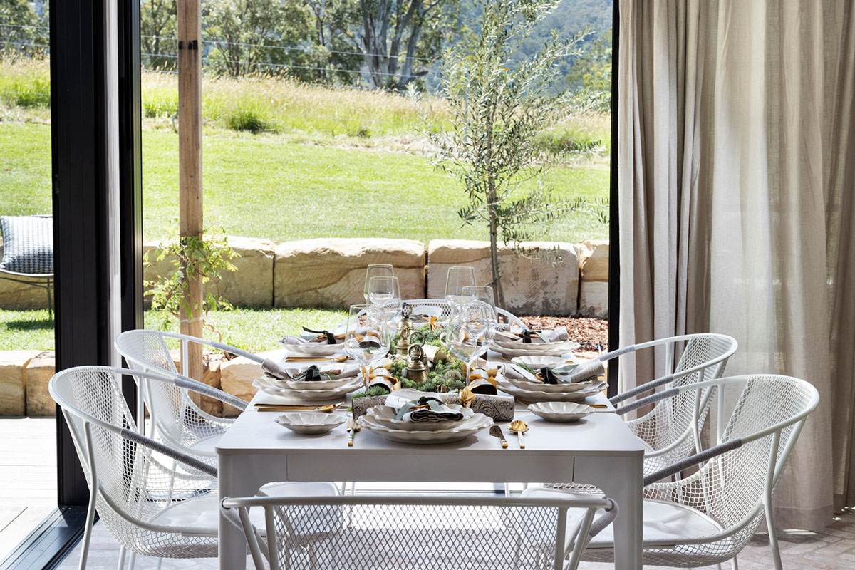 A white dining table and chairs set for a festive meal, featuring layered place settings and natural light from large windows overlooking a garden.