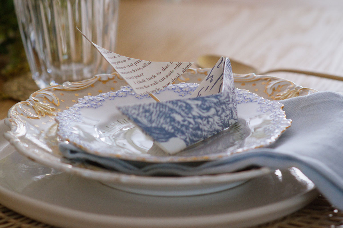A place setting with gold, white and blue China tableware, topped with a handcrafted origami boat.