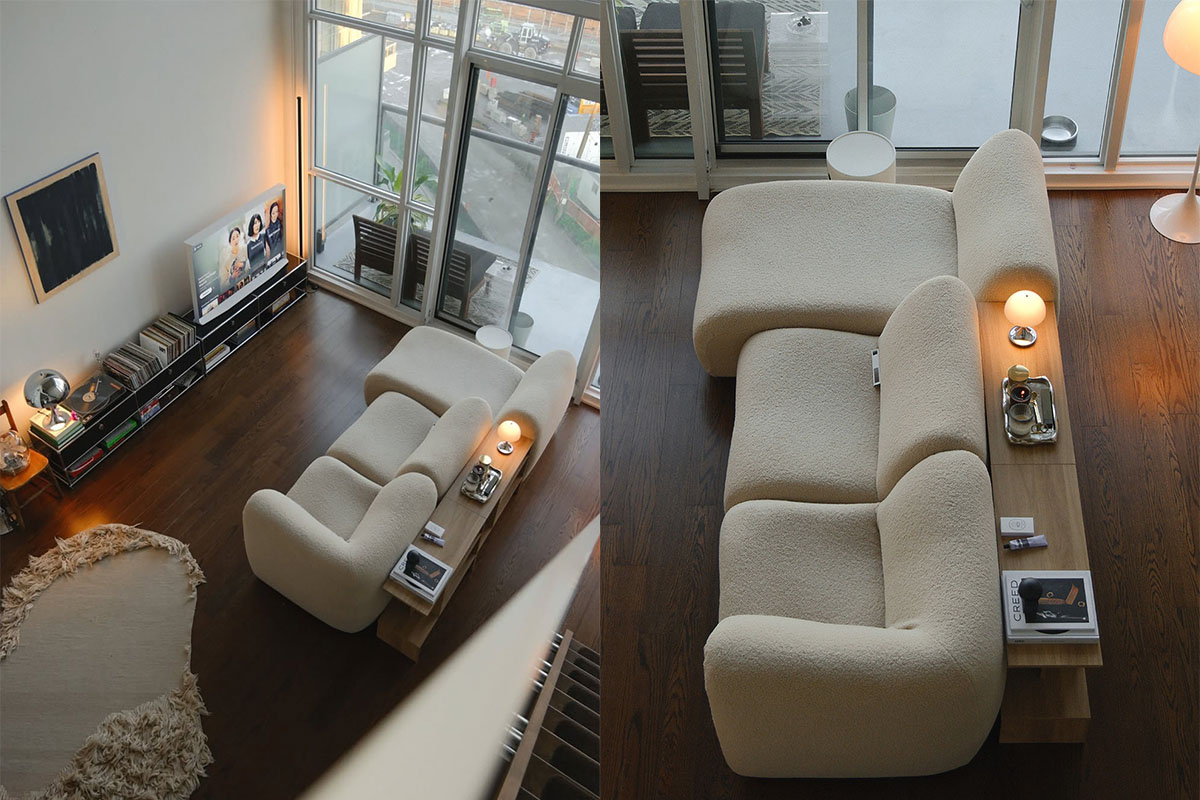 Aerial views of a curved ivory modular sofa with timber console, styled in a compact loft apartment with dark timber flooring, full-height windows and a warm, minimalist aesthetic.