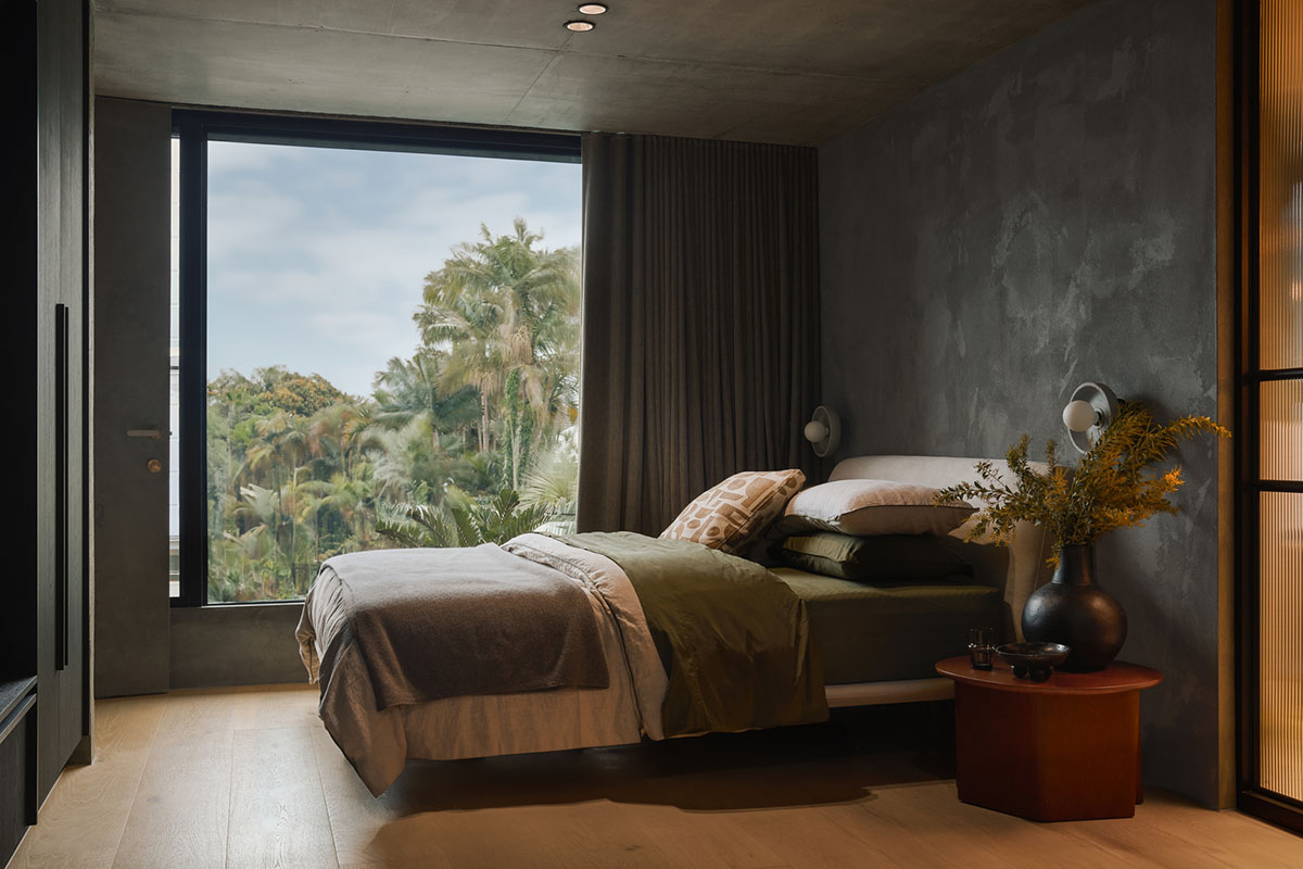 Guest bedroom with a queen bed, layered bedding in soft neutrals and green, dark textured walls and a window framing lush greenery.
