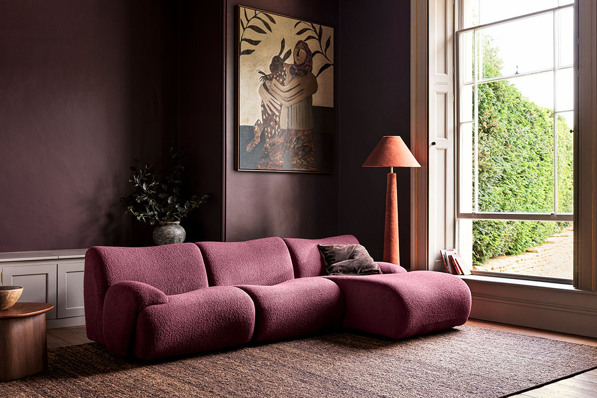 Deep plum modern sofa in a living room with high ceilings, artwork and a wide window overlooking trees.
