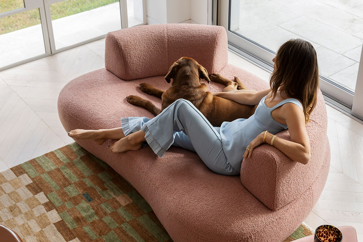 Kate lounges with her dog on a curved, textured dusky pink lounge chair by a sunlit window.