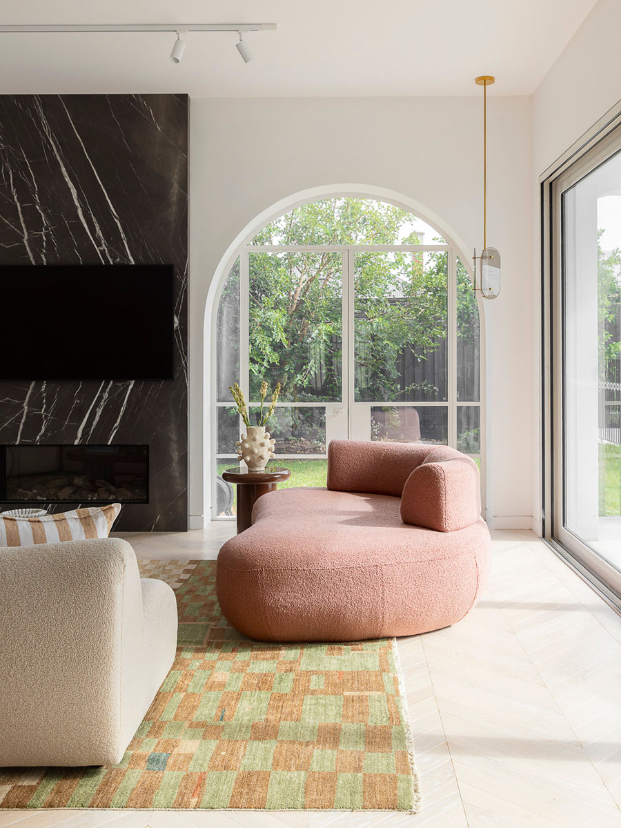 A softly curved pink sofa sits in contrast to the structured lines of a checkered rug, built-in joinery and an arched window framing a lush garden view.