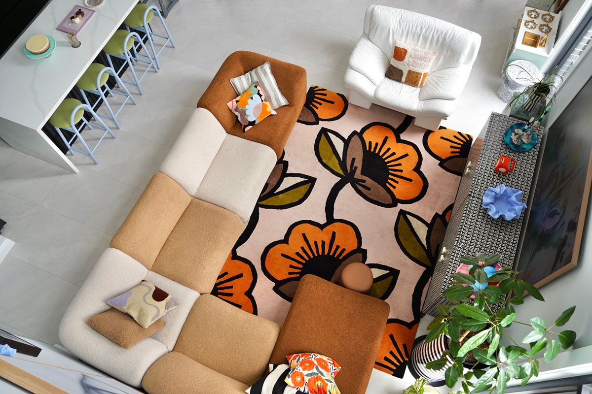 Overhead shot of a living and dining room, featuring a large, curved, modular sofa in a warm, neutral palette of caramel and beige tones, styled with a bold floral rug and eclectic accents.