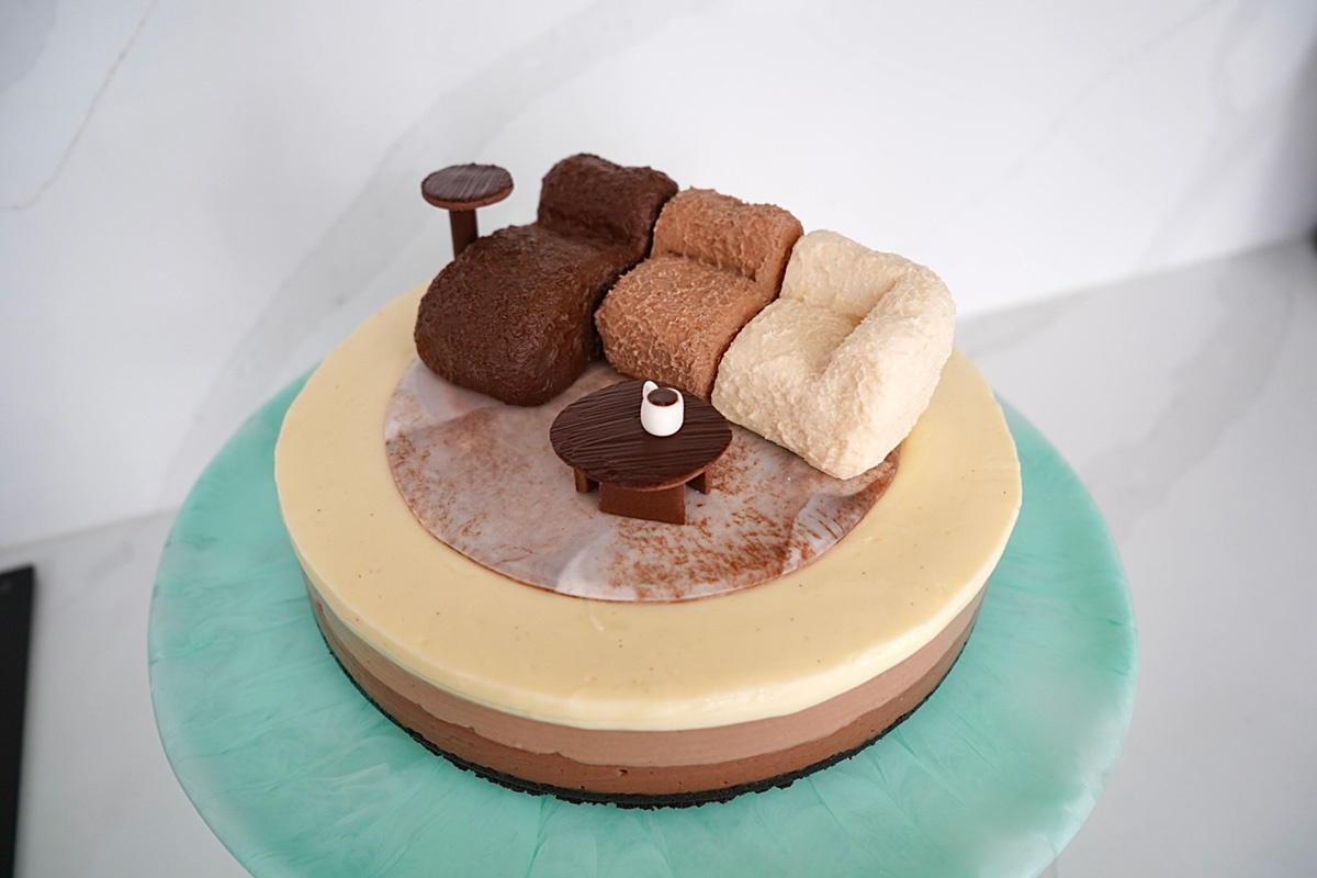 Wide shot of Katherine Sabbath’s 1997 Sofa inspired mousse cheesecake, displaying three layers of dark, milk and white chocolate with edible side table and coffee cup accents.