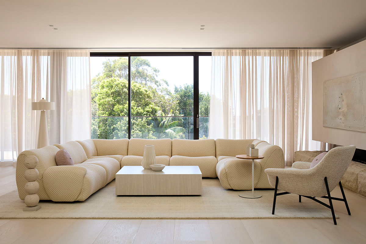 A wide view of Lara Hutton’s light-filled modern living space, featuring a curved modular sofa, rectangular coffee table and an upholstered armchair. Lush outdoor greenery frames tonal King Living designs.