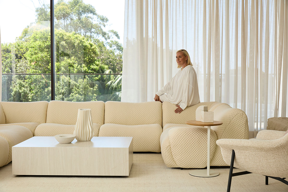 Interior Designer Lara Hutton sits on a large, cream modular sofa in a contemporary, light-filled living room with minimalist furniture.