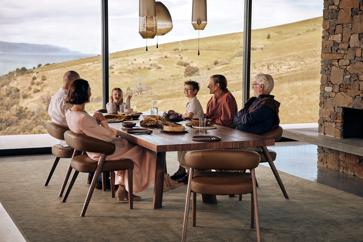 Family gather around a timber dining table in a modern dining room with hilltop views.