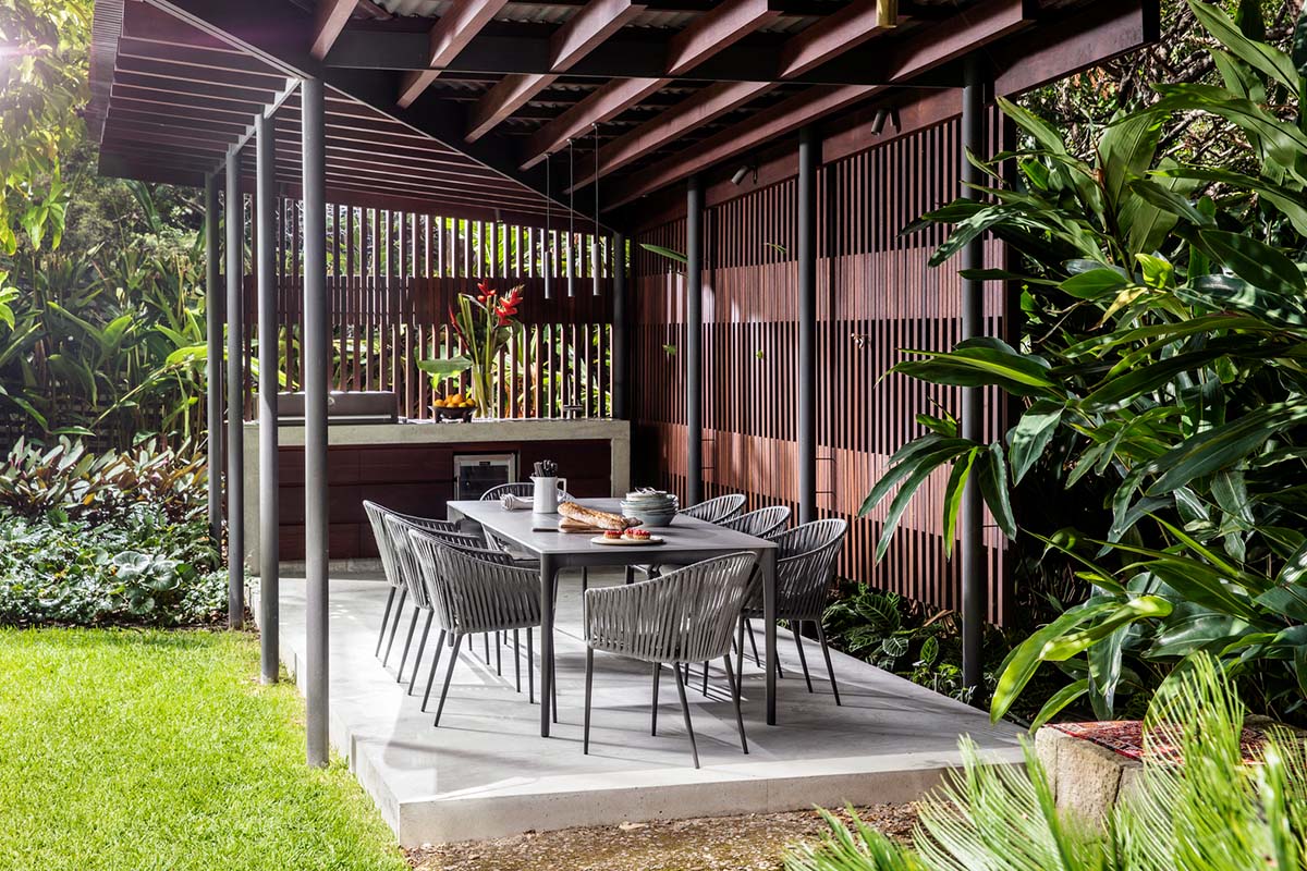 Outdoor dining setting with woven chairs and ceramic table under covered pergola surrounded by lush greenery.