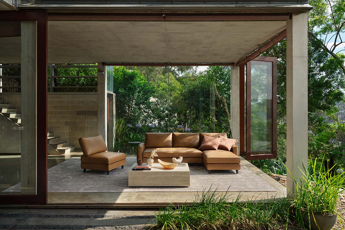 Brown leather sofa in a modern living room with large glass windows overlooking forest surroundings.