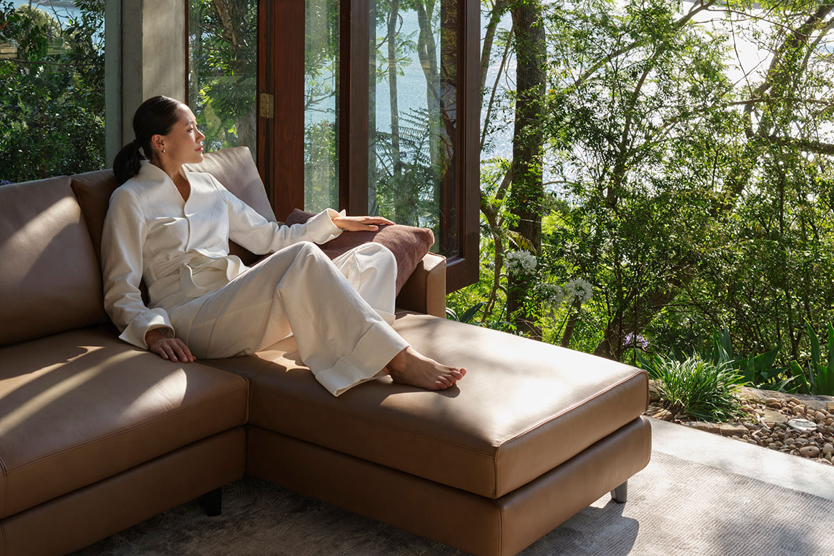 Woman dressed in white rests on a brown leather sofa overlooking trees.
