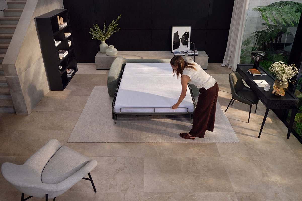 Modern living space with a woman unfolding a sleek sofa bed, surrounded by contemporary decor and greenery.