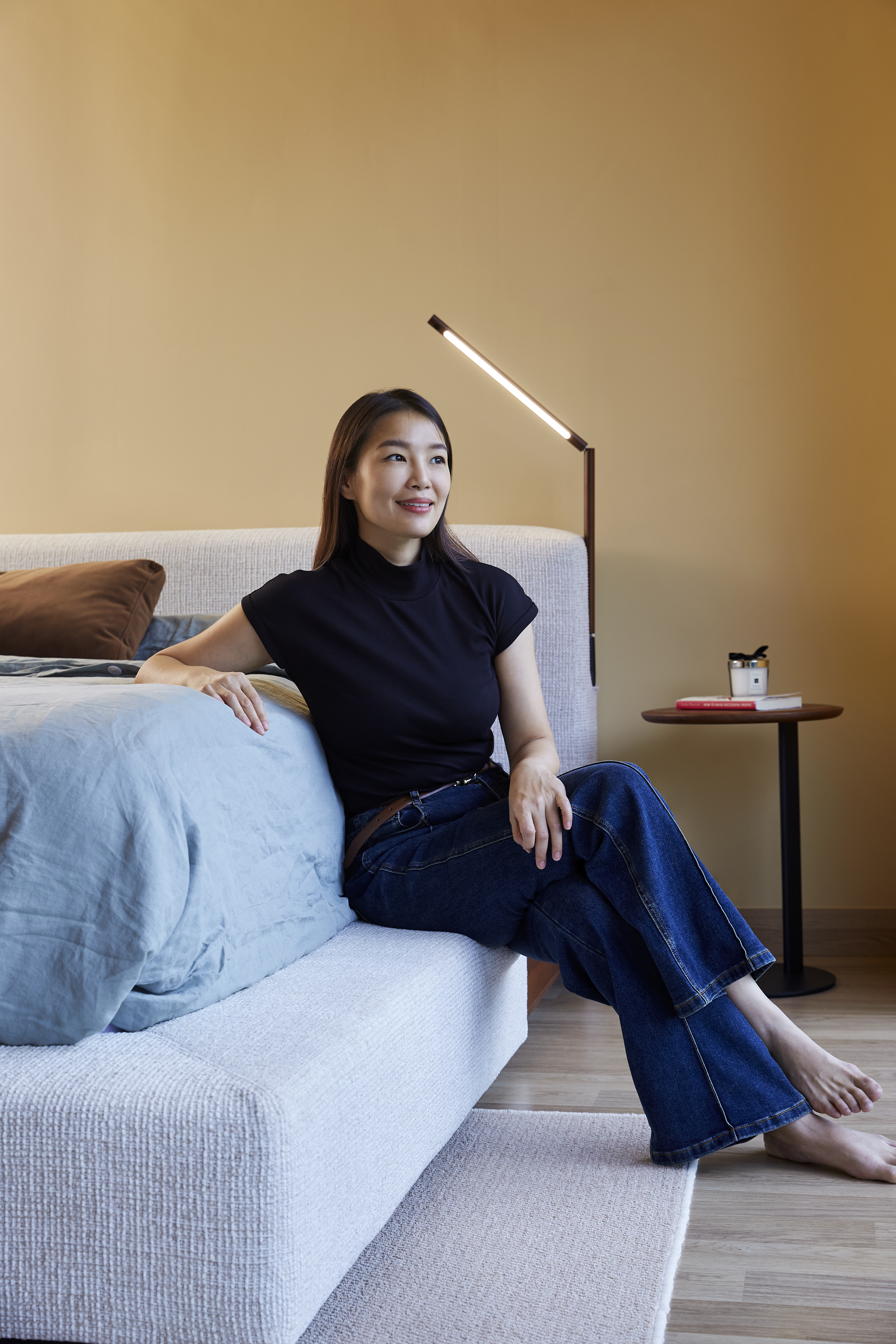 Person sitting on a modern upholstered bed with a minimalist lamp and side table in the background.