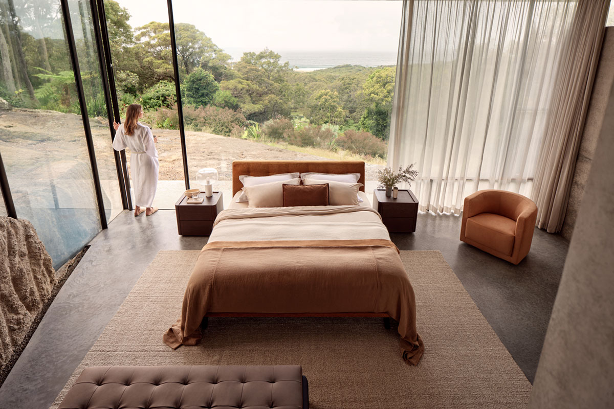 A warm, autumnal bedroom with a burnt orange bed styled centrally, surrounded by a tonal armchair, deep timber bedside tables and a chocolate brown leather ottoman.
