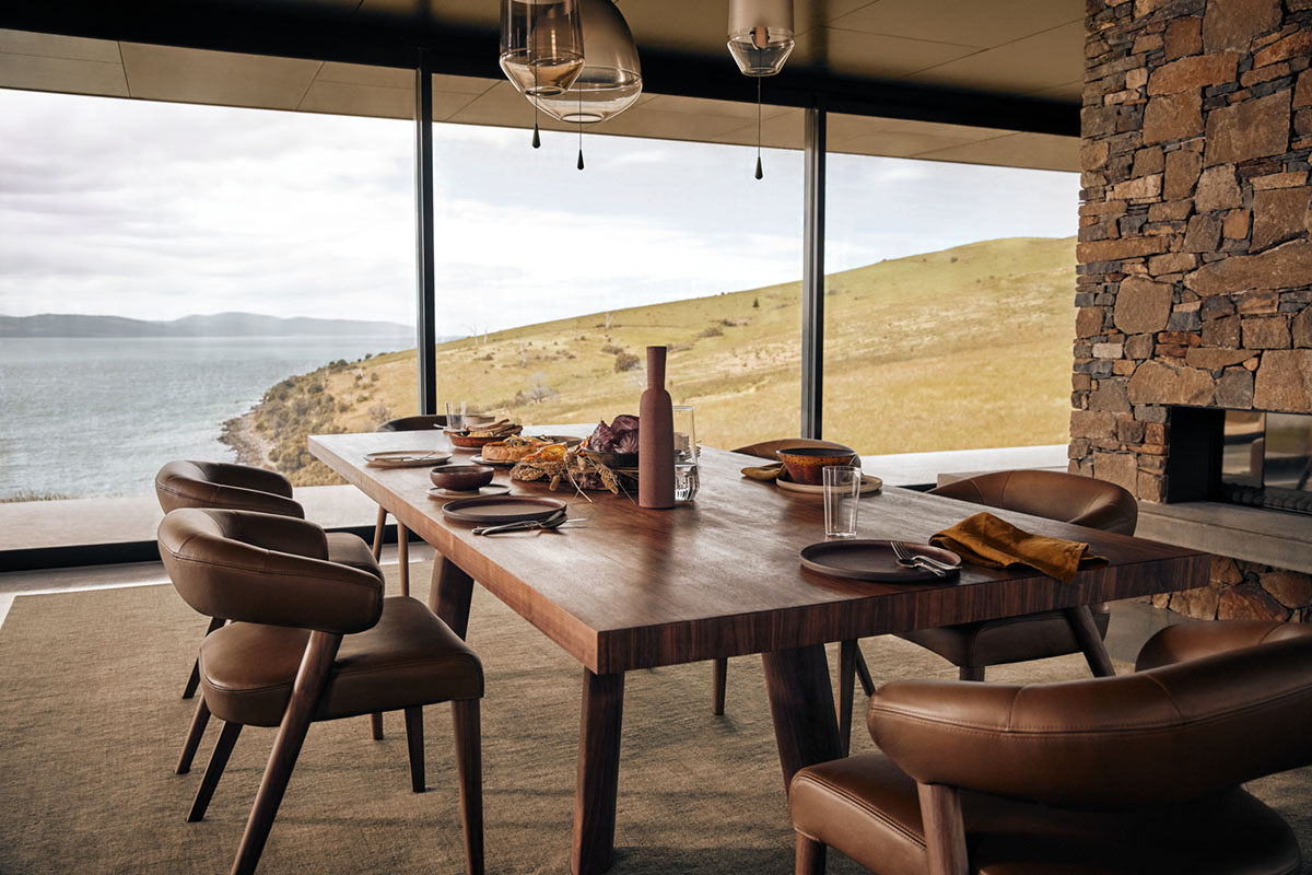 Dining setting with rich timber table and brown leather chairs styled with warm-toned tableware, framed by a brick fireplace and expansive coastal views.