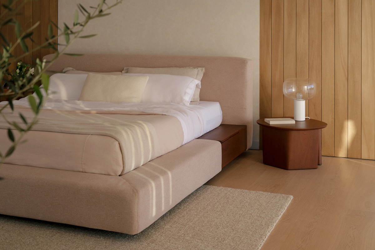 A fabric bed in soft pastel pink with integrated timber shelving for storage in a neutral bedroom styled with florals.